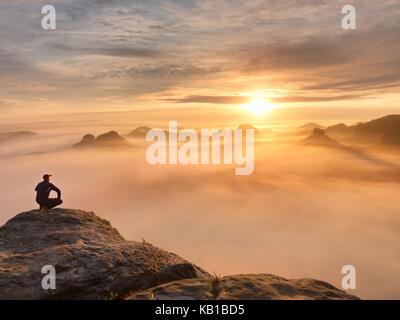 Tourist sit on peak of sandstone rock and watching into colorful mist and fog in  morning valley. Sad man. Man sit. Man in jeans.Fall mountain mist. M Stock Photo