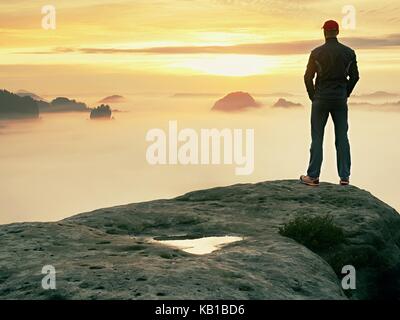 Man stands alone on the peak of rock. Hiker watching to autumn  Sun at horizon . Beautiful moment the miracle of nature. Colorful mist in valley. Man  Stock Photo