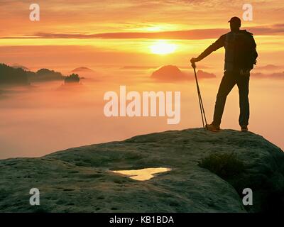 Man silhouette with poles in hand. Sunny spring daybreak and tourist guide stay on sharp cliff of mountain. Hiker with sporty backpack stand on rocky  Stock Photo