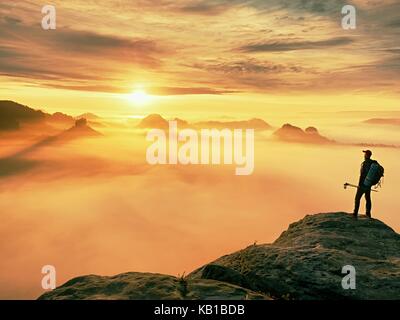 Man silhouette with poles in hand. Sunny spring daybreak and tourist guide stay on sharp cliff of mountain. Hiker with sporty backpack stand on rocky  Stock Photo