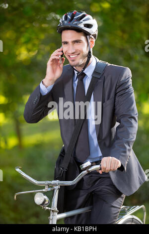 young businessman wearing bike helmet, riding bicycle and using mobile phone, outdoors Stock Photo
