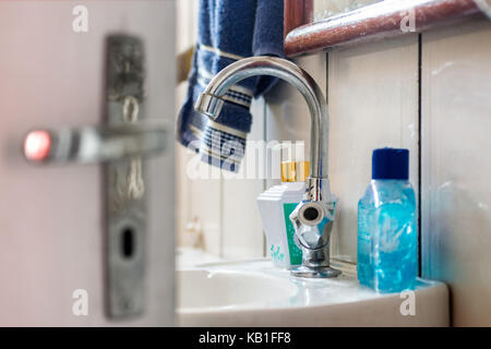 Interior of a bathroom, showing white tiled walls, door in the foreground with lock and handle, a white sink with metal faucet, liquid soap, mirror Stock Photo