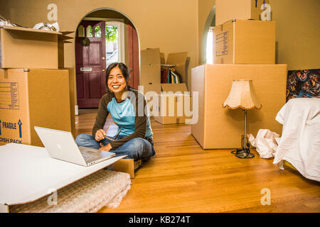 A portrait of a young Asian woman sitting on the floor in front of a laptop computer surrounded by moving boxes as if she is moving in or out. Stock Photo