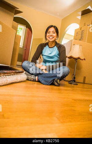 A portrait of a young Asian woman sitting on the floor surrounded by moving boxes as if she is moving in or out. Stock Photo