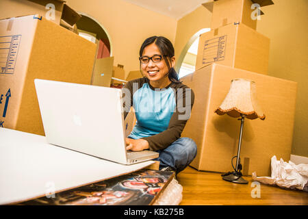 A smiling young woman sitting on the floor surrounded by moving boxes working on a laptop computer. Stock Photo