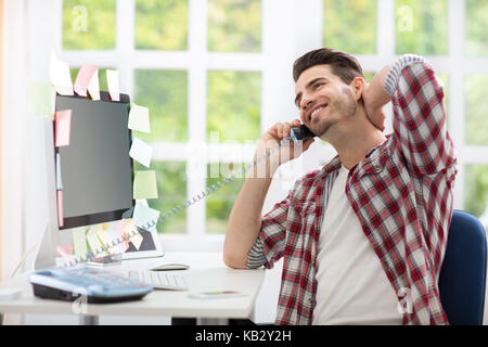 Cheerful young man sitting at work desk front of computer and  talking on phone Stock Photo