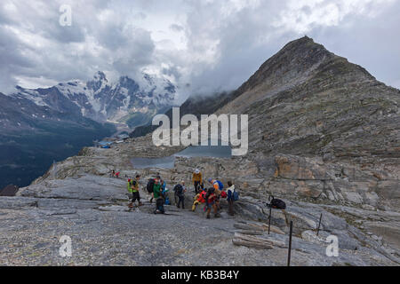 MORO PASS NEAR MACUGNAGA, ITALY - JULY 2017; Group of hikers and Smeraldo lake with Rosa Mount on the background. Stock Photo