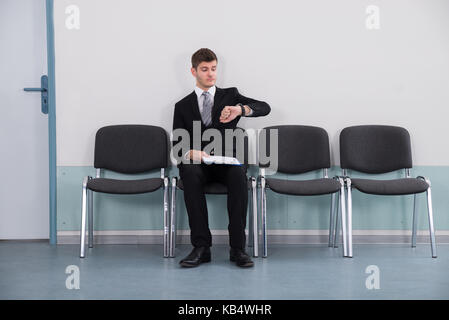 Young Businessman Sitting On Chair Looking At Watch Stock Photo