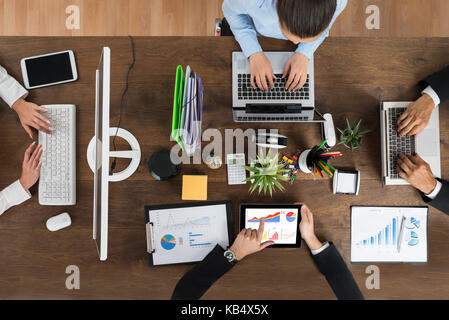 High Angle View Of Businesspeople Working On Electronic Devices At Wooden Desk Stock Photo