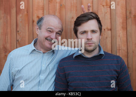 Father pranking his son with bunny ears isolated on wooden background. Young nman is very serious, old gentleman smiles. Stock Photo