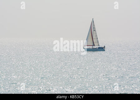 Small yacht on the sea with sun sparkling in the water. Small sailing boat on the ocean with reflecting sun sparkles. Stock Photo