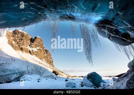 View from ice cave towards sunlit mountains with icicles hanging from cave entrance, near Jokulsarlon, South Icelan Stock Photo