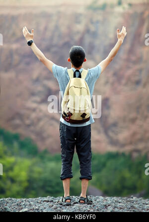 Young tourist man with backpack standing with raised hands on rocky cliff Stock Photo