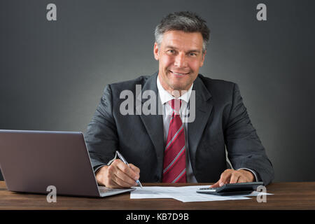 Mature businessman checking bills with calculator and laptop at desk against gray background Stock Photo