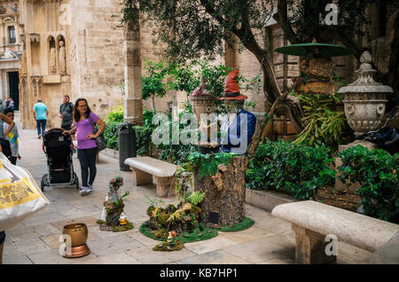 Valencia, Spain - June 3, 2017: Living statue of a man painted in vibrant colors and seated on stump with fountain on street. Stock Photo
