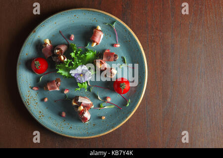 Spanish jamon twisted in roll with dates, decorated with cherry tomato and pomegranate. Top view of plate with prosciutto. Stock Photo