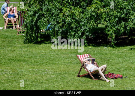 A woman reading a book and enjoying the warm sunshine and hot weather is pictured sitting in a deckchair in a public park in Bath Somerset England UK Stock Photo