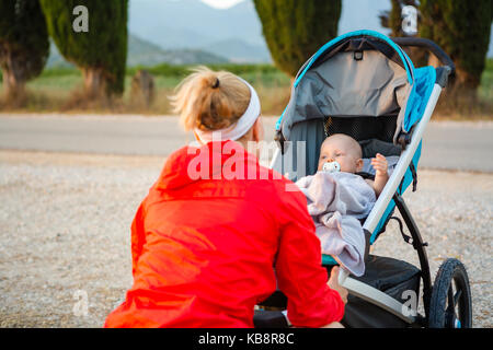 Mother with child in stroller enjoying motherhood at sunset and mountains landscape. Jogging or power walking woman with pram at sunset. Beautiful ins Stock Photo