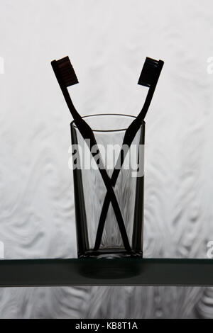 Two toothbrushes facing each other in a glass, silhouetted against a textured bathroom window. Shallow depth of field with focus on glass rim. Stock Photo