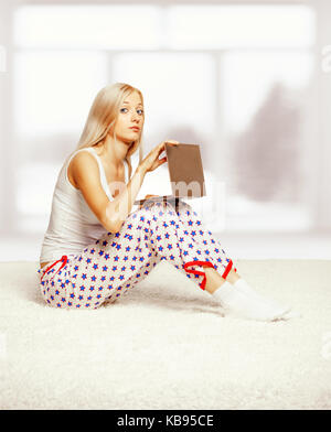 Young blonde woman in pyjamas on white whole-floor carpet browsing laptop  near window Stock Photo