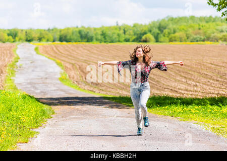 Young woman running, jumping in air and smiling on countryside dirt road by brown plowed fields with furrows in summer in Ile D'Orleans, Quebec, Canad
