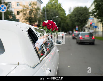 Unidentified bride stuck out her hand with the red bouquet from the window of the decorated white car and lightly waving it on warm summer day. Stock Photo