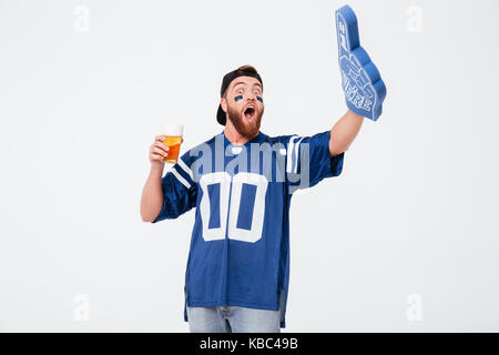 Picture of emotional man fan in blue t-shirt wearing fan finger number one glove standing isolated over white background. Looking aside drinking beer. Stock Photo