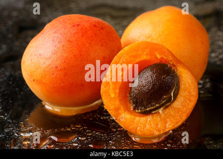 Close up of two whole and one half sliced apricot fruits with the stone, ripe, freshly washed with water drops on a dark background Stock Photo