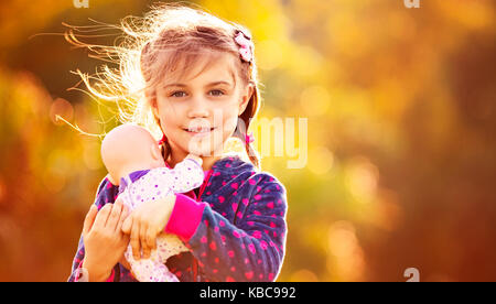 Portrait of a sweet baby girl with doll over orange autumnal foliage background, happy child playing mother's daughters outdoors in fall sunny day Stock Photo