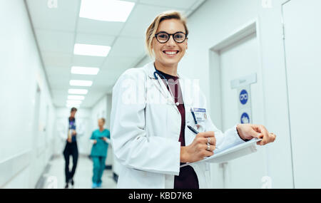 Portrait of happy young female physician standing in corridor with clipboard. Caucasian woman working in hospital writing prescription. Stock Photo