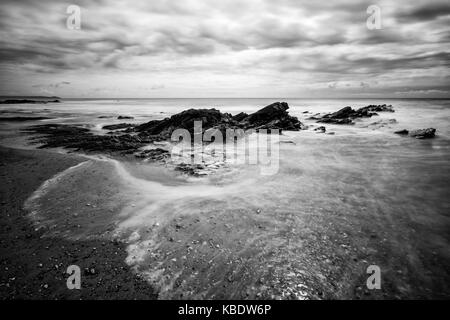 Jagged rocks and soft water lapping upon the beach. This image was taken at Seaton, Cornwall, UK. Stock Photo