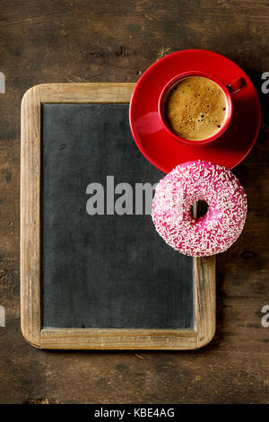 Empty vintage chalkboard, pink glazed donut and red cup of black coffee over old wooden background. Top view with space for text. Stock Photo