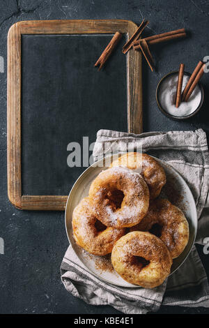 Plate of homemade donuts with sugar and cinnamon powder on vintage chalkboard served with spices and textile napkin over dark texture background. Top  Stock Photo