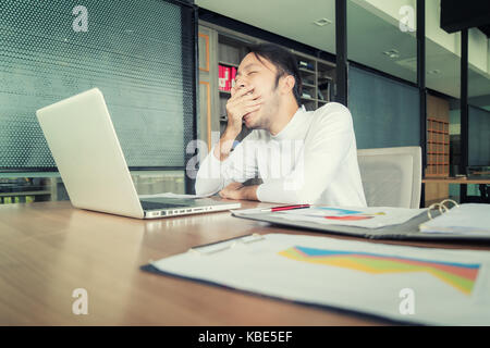 Asian tired businessman in casual wear yawning after long working day at workplace in office. Stock Photo