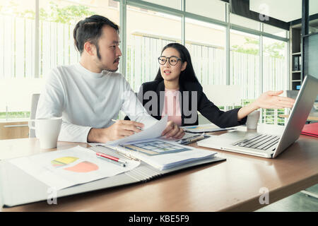 Couple of young designers working at modern office, two coworkers discussing fun project over a laptop, little team of businesspeople smiling and look Stock Photo
