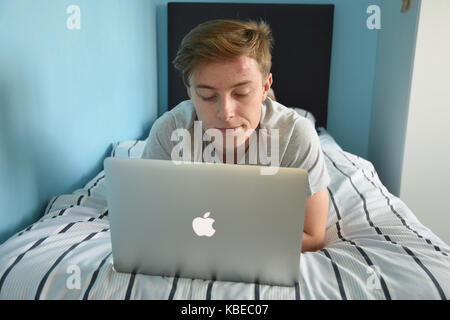 19 year old teenage boy reading from a Apple Macbook Air laptop in his bedroom Stock Photo