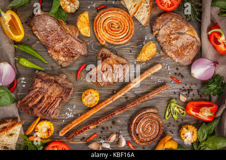 Assorted delicious grilled meat and sausages with vegetables on a wooden background. Still life - delicious roasted pieces of meat and sausages with p Stock Photo