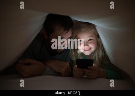 Father and daughter lying under blanket and using mobilephone at home Stock Photo