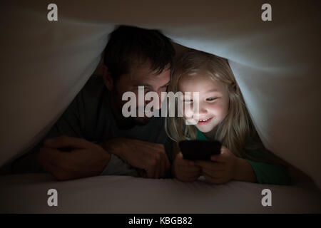 Father and daughter lying under blanket and using mobilephone at home Stock Photo