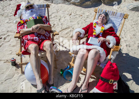 Santa Claus in red swimming trunks and Hawaiian shirt lounging on sandy beach with Mrs Claus sleeping on sandy beach. Stock Photo