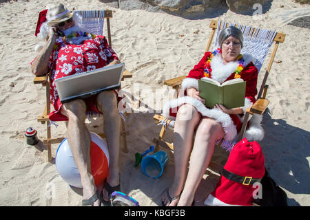 Santa Claus in red swimming trunks and Hawaiian shirt working on laptop computer on sandy beach with Mrs Claus talking on cell phone. Stock Photo