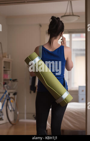Rear view of young woman with exercise mat talking on phone at home Stock Photo
