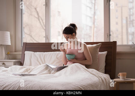 Young woman having fruit while reading book on bed at home Stock Photo