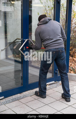 Hooded man using crowbar to open glass door Stock Photo
