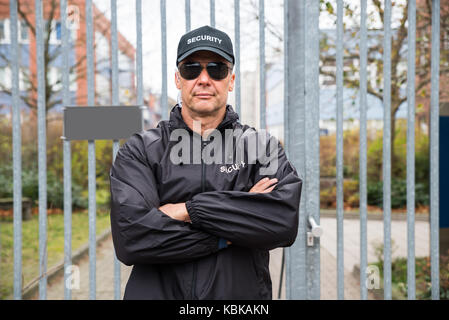 Portrait of confident mature security guard standing arms crossed in front of gate Stock Photo