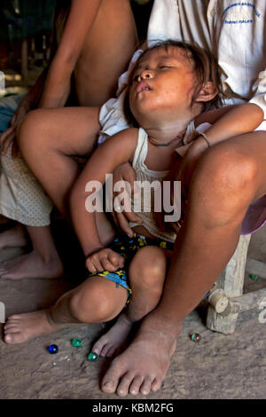 A poor & exhausted young Cambodian girl is sitting on the dirt floor of a home while a boy holds her in a village in Kampong Cham Province, Cambodia.. Stock Photo