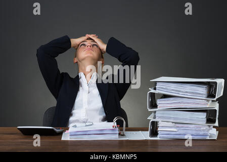 Tired businesswoman with hands on head sitting at desk over gray background Stock Photo