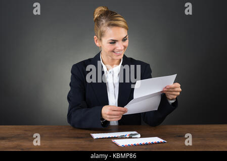 Smiling young businesswoman reading letter at desk while sitting over gray background Stock Photo