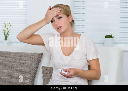 Young woman holding thermometer while touching head at home Stock Photo