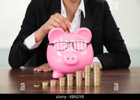 Close-up Of Businesswoman Inserting Coin In Piggybank In Front Of Coins Stack Stock Photo